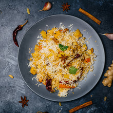 Load image into Gallery viewer, Image of chicken biryani in a large bowl istting on a stone surface. Surrounded by various fresh spices such as chilli, cinnamon, garlic and ginger 
