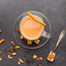 Load image into Gallery viewer, Image of a glass tea cup and saucer containing masala chai. Surrounded by fresh spices and a metal spoon with some chai masala
