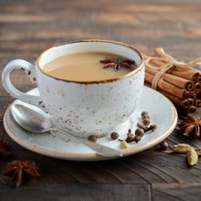 Load image into Gallery viewer, Image of a ceramic tea cup and saucer containing masala chai. With a teaspoon sitting on the saucer and surrounded by fresh spices
