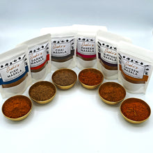 Load image into Gallery viewer, Image of the full range of Sashi&#39;s masalas. 6 packets of 50g masalas and each packet has a gold metal bowl sitting in front containing the respect masala. 
