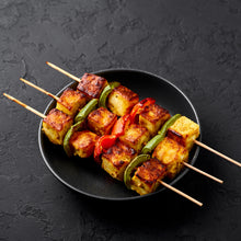 Load image into Gallery viewer, Image of some grilled paneer on skewers wiht peppers, on top of a black bowl which is sitting on a black stone surface
